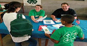 photo of seated volunteers chatting with standing 4-H youth at a table with 4-H-branded materials.
