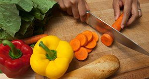 close up of a person slicing a carrot on a cutting board with vegetables and bread.