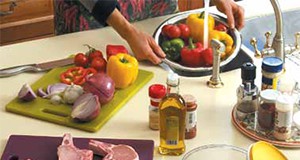 a photo of a food preparation area in a home kitchen, with a person rinsing vegetables at the sink, separate cutting boards for vegetables and meat, and an array of ingredients at hand.