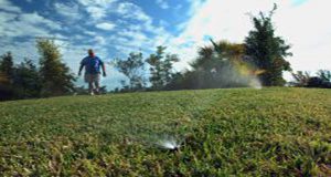 close up photo of a lawn irrigation sprinkler head with a lawn, person walking and trees in the background