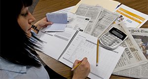 woman writing in financial ledger with newspapers, check register, and calculator at hand