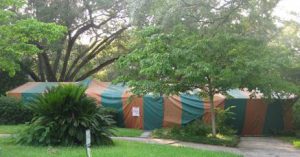 A modest Florida ranch house with dogwoods, oak trees, and a largec cycad in the yard. House is completely covered by a green and orange striped tent with a warning signed pinned near the front walkway indicating fumigation.