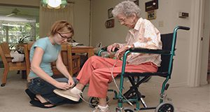 A caretaker kneeling in front of an elderly woman in a wheel chair and tying her shoe. 