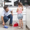 A father crouching and holding a dustpan while his young daughter sweeps the kitchen.