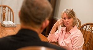 A woman holding a tissue sitting at a table talking with a man with his back to the camera.