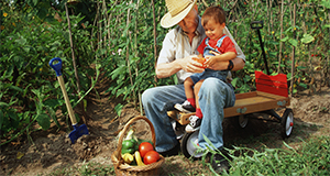 Grandparent and grandchild sitting on a wagon in a field near a basket of vegetables.